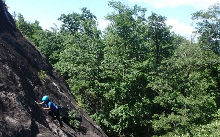 A student wearing safety gear climbs up a rock incline in front of green trees.
